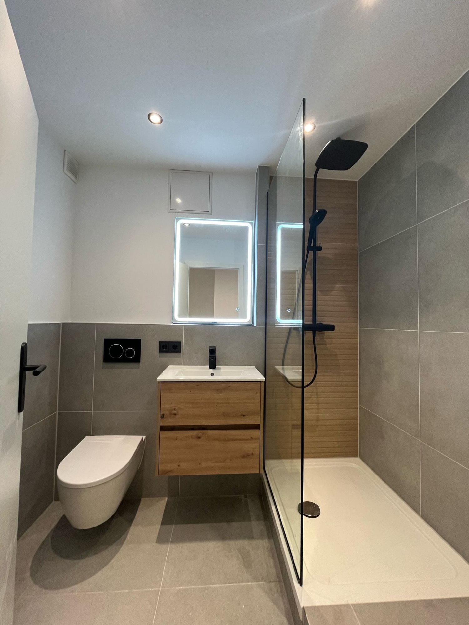 Modernisiertes Bad mit bodengleicher Dusche und Glasabtrennung, wandhängendem WC, Holzwaschtisch, LED-Spiegel und grauen Großformatfliesen